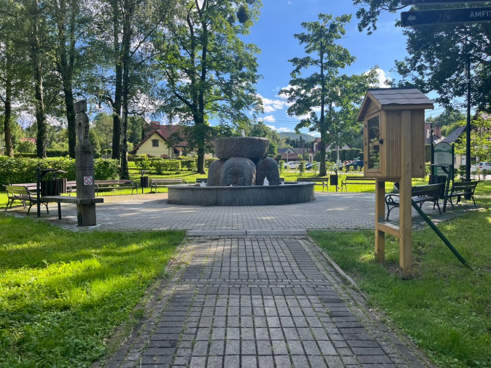 Plac z kostki brukowej wokół kamiennej fontanny w Parku Zdrojowym w Jaworzu, ławki i tablice informacyjne.