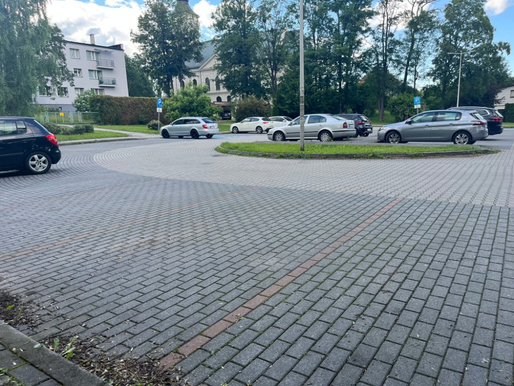 Parking przy parku z zaparkowanymi samochodami, chodnik z kostki i przejście, w tle zabudowania.