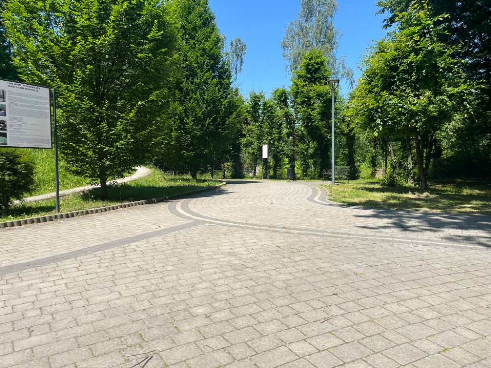 Rozległy brukowany plac z tablicą informacyjną, latarnią i młodymi drzewami pod błękitnym niebem.