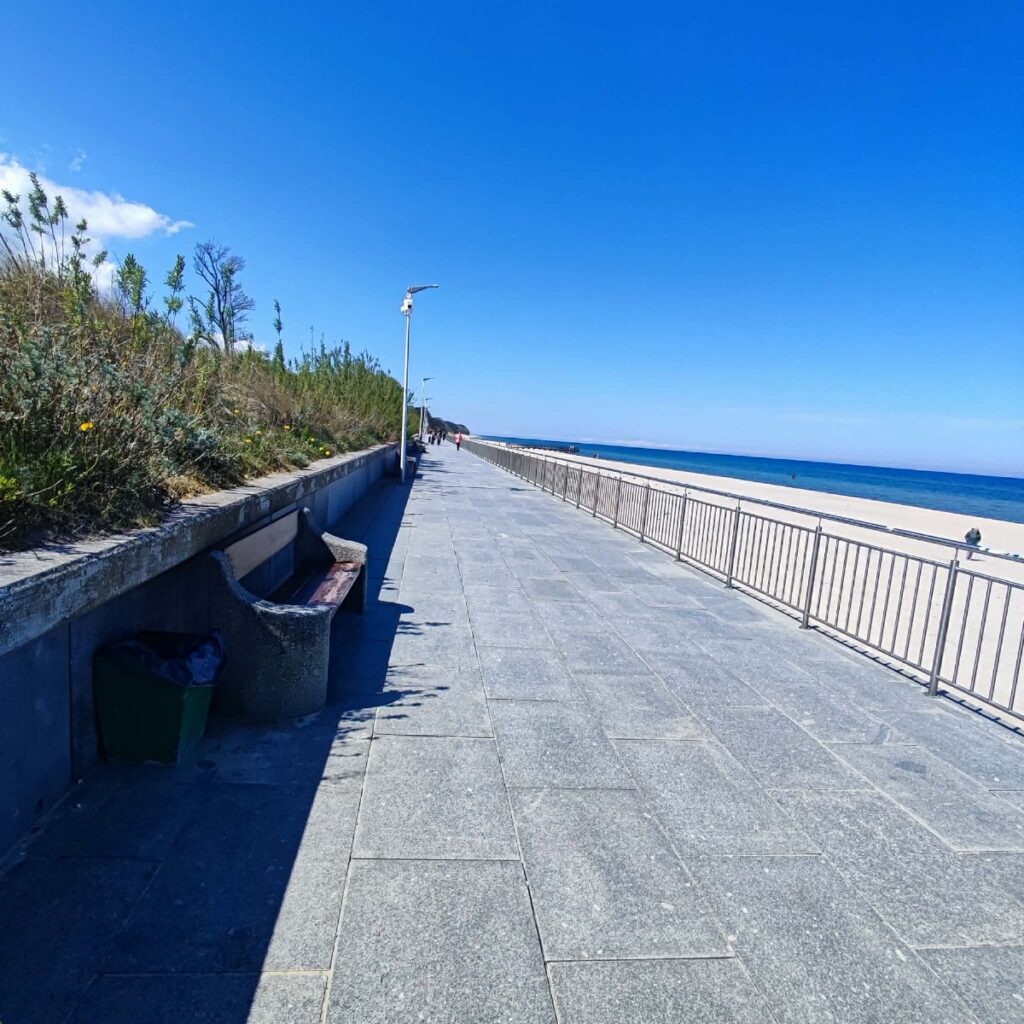 Szeroki granitowy deptak promenady w Sarbinowie z ławką i koszem na śmieci, po prawej barierka oddzielająca plażę i morze.