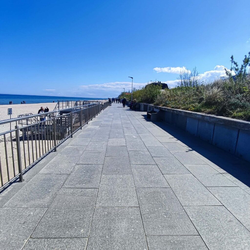 Promenada nadmorska w Sarbinowie z równą nawierzchnią z płyt granitowych, spacerowicze, po lewej zejście na plażę z poręczami.