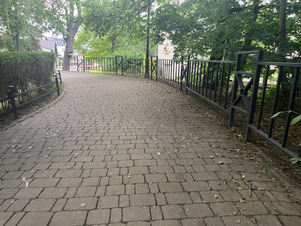 Alejka z kostki biegnąca wzdłuż metalowej barierki nad wodą, przy żywopłocie.