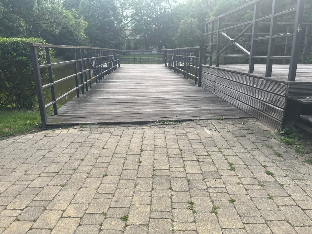 Drewniany most z barierkami, dojście z kostki brukowej, po prawej schody.