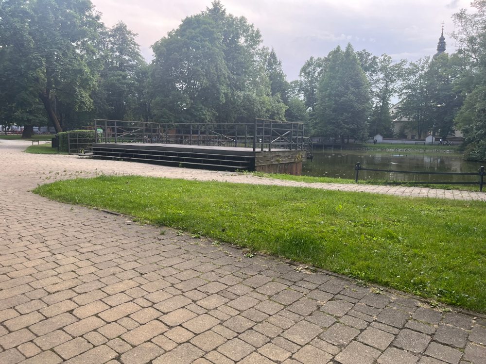 Alejka z kostki brukowej nad stawem, w tle zadaszona scena amfiteatru i trawnik.