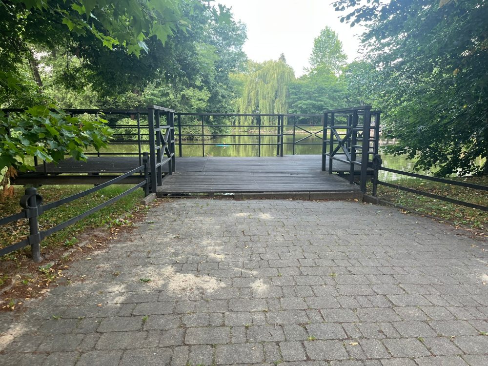 Chodnik z kostki prowadzący na drewniany mostek z metalowymi barierkami nad stawem.