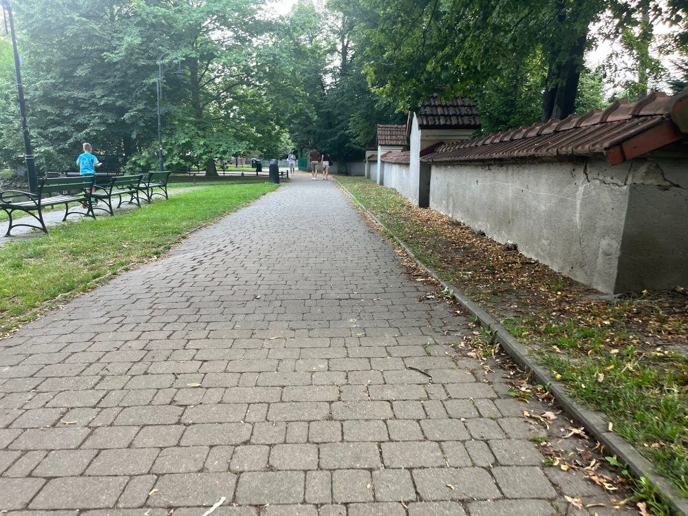 Szeroka alejka z kostki wzdłuż murowanego ogrodzenia krytego dachówką, po lewej ławki.