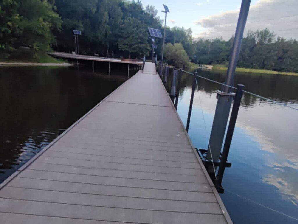 Długa, prosta kładka z desek biegnąca nad wodą, po prawej metalowe słupki, w tle las.