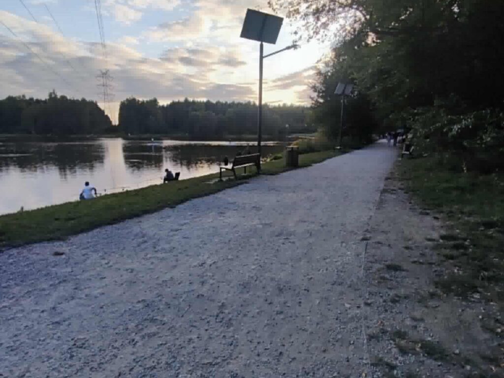 Żwirowa alejka wzdłuż brzegu zbiornika, ławka i lampa solarna, ludzie nad wodą.