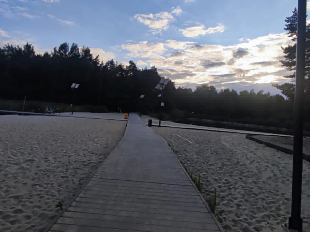 Utwardzona ścieżka wzdłuż piaszczystej plaży, lampy solarne, zmierzch nad lasem.
