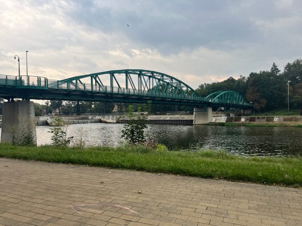 Brukowana promenada nad brzegiem Odry, za nią trawnik i zielony most kratownicowy.