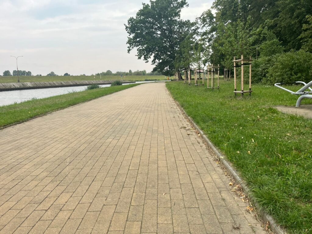 Szeroka alejka z kostki wzdłuż betonowego koryta rzeki, obok młode drzewa i metalowa leżanka.