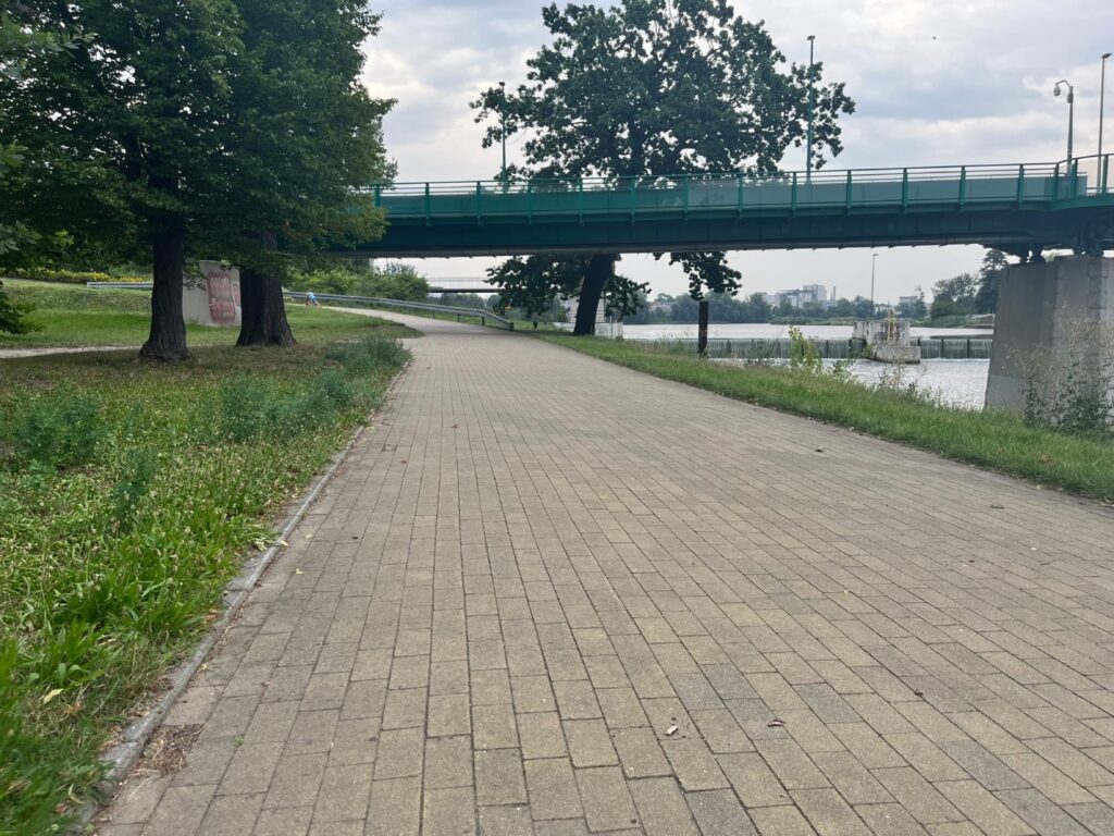 Brukowana alejka przechodząca pod mostem drogowym w jednym poziomie, bez schodów i progów.