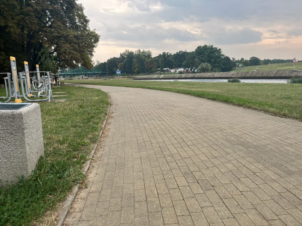 Równa nawierzchnia z kostki przy siłowni zewnętrznej na betonowych podestach, obok trawnik i rzeka.