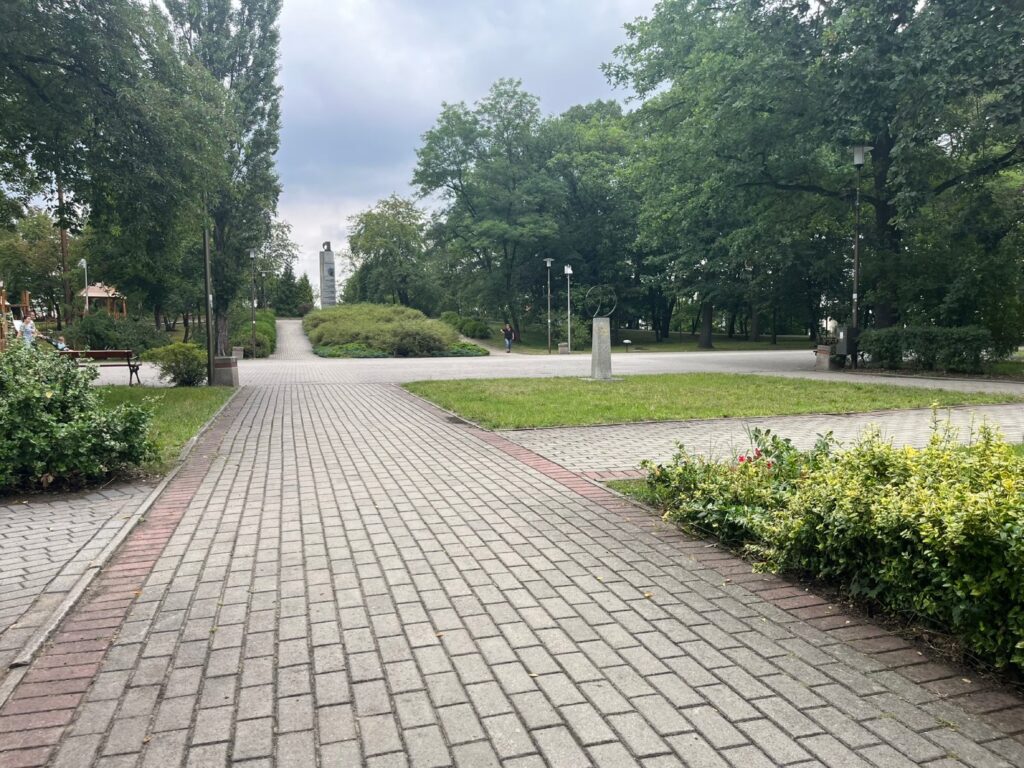 Skrzyżowanie brukowanych alejek, w głębi pomnik Pojednania na wzniesieniu, po lewej plac zabaw, po prawej rzeźba na cokole.