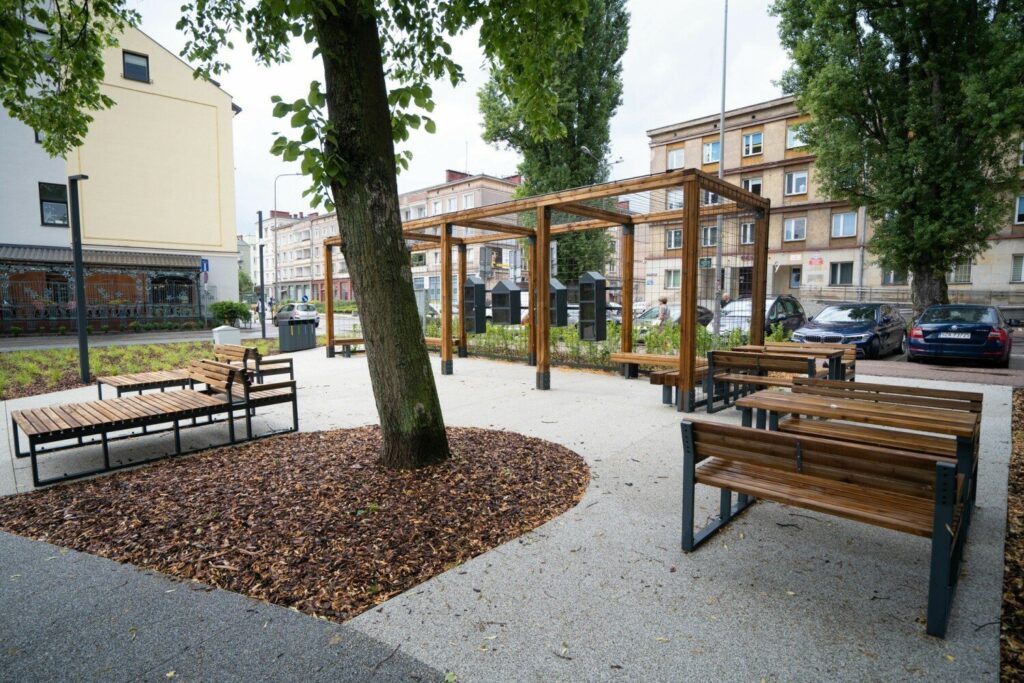 Drewniana pergola z ławkami nad jasną, ubitą nawierzchnią mineralną, w tle kamienice w centrum Kielc.
