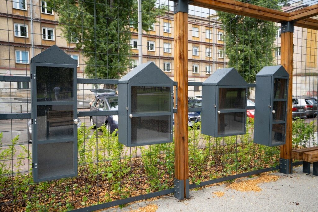 Cztery przeszklone gablotki pawilonu wymiany książek zamocowane na słupach drewnianej pergoli.