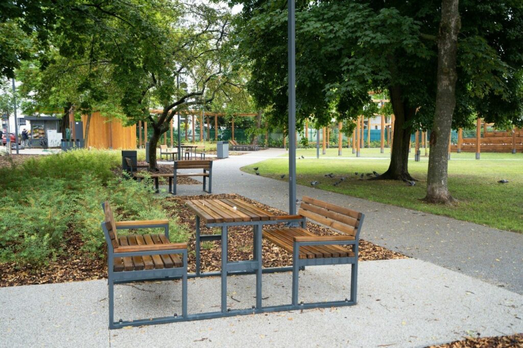 Zestaw ławek ze stolikiem przy alejce, dalej trawnik, stare drzewa i ogrodzony plac zabaw.