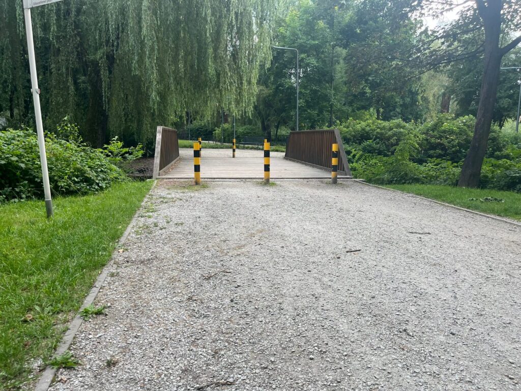 Dojazd z luźnego grysu do kładki z metalowymi barierkami, na wjeździe żółto-czarne słupki.