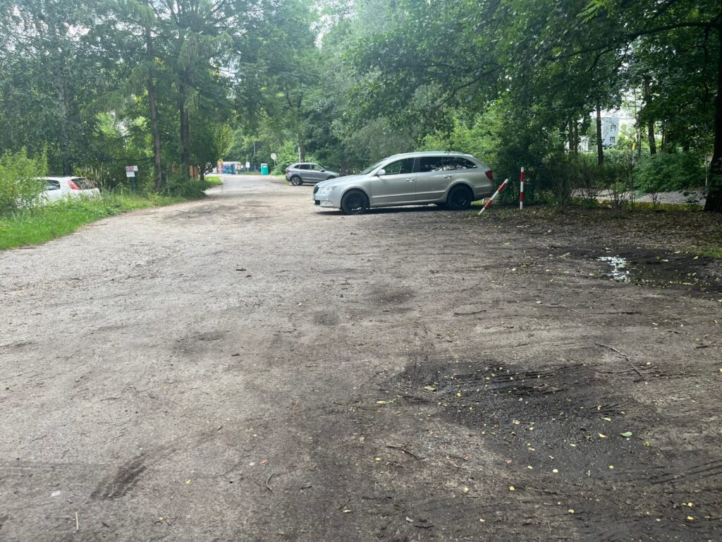 Nieutwardzony plac parkingowy z kałużami i koleinami, samochody zaparkowane pod drzewami.