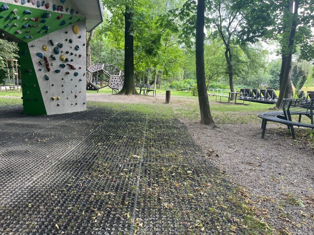 Ścianka bulderingowa na ażurowych gumowych matach, w tle kopuła wspinaczkowa, ławki i leżaki pod drzewami.