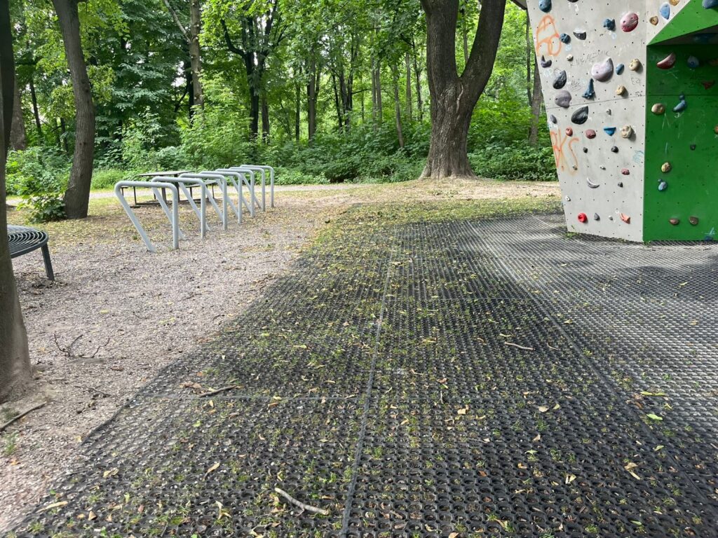 Gumowe maty pod ścianką wspinaczkową, obok ubity plac z metalowymi stojakami rowerowymi.