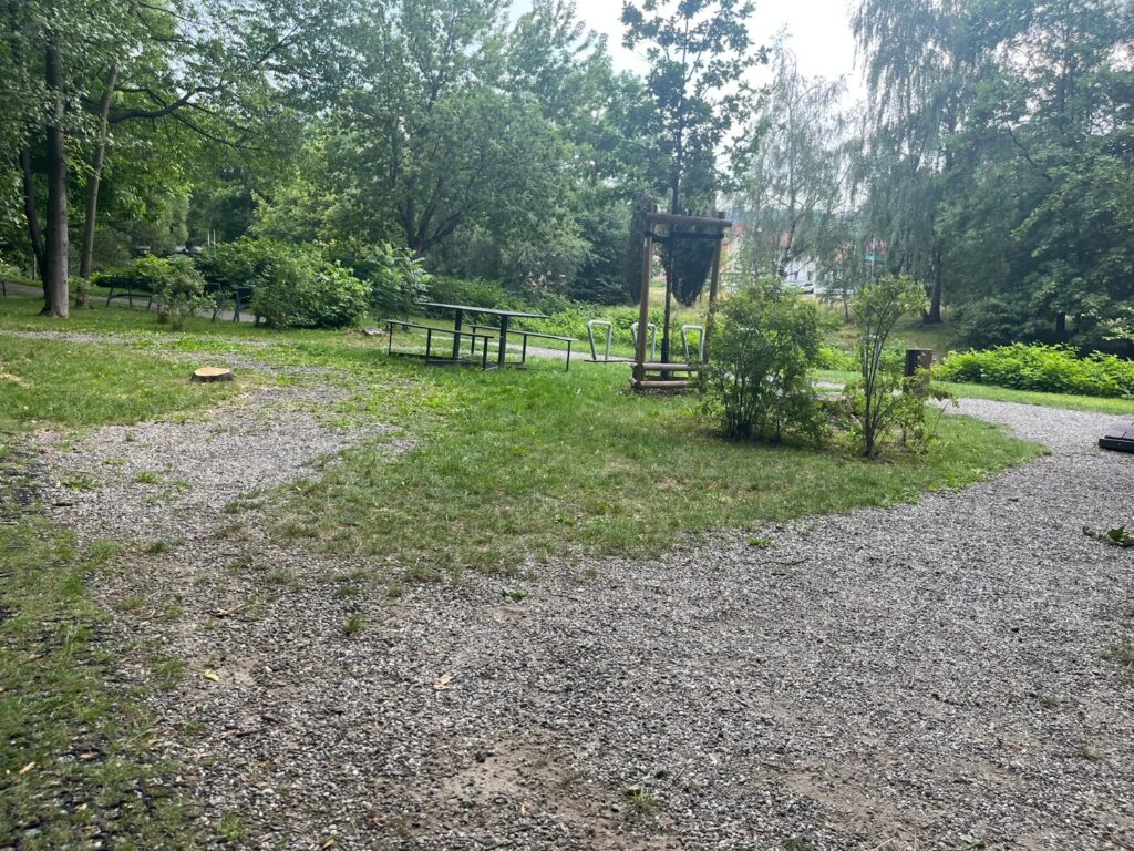 Ścieżka z luźnego grysu wśród trawnika, dalej stół piknikowy z ławkami i drewniana konstrukcja.