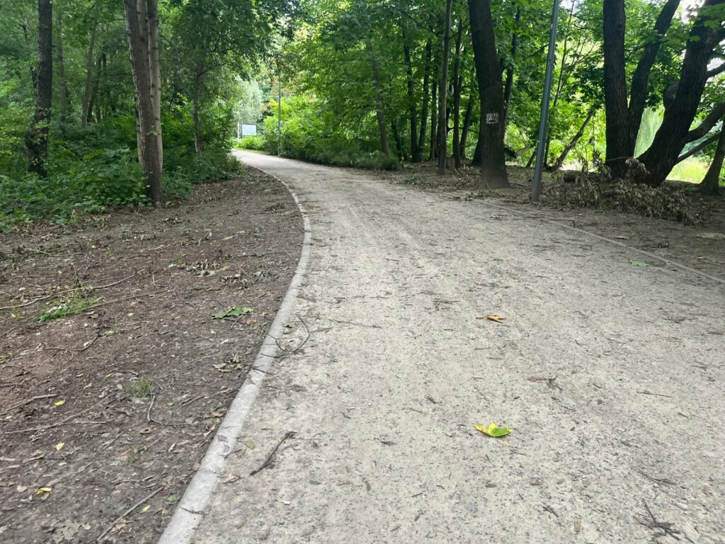 Łagodny zakręt szerokiej alei szutrowej z betonowym obrzeżem, po bokach las i latarnie.