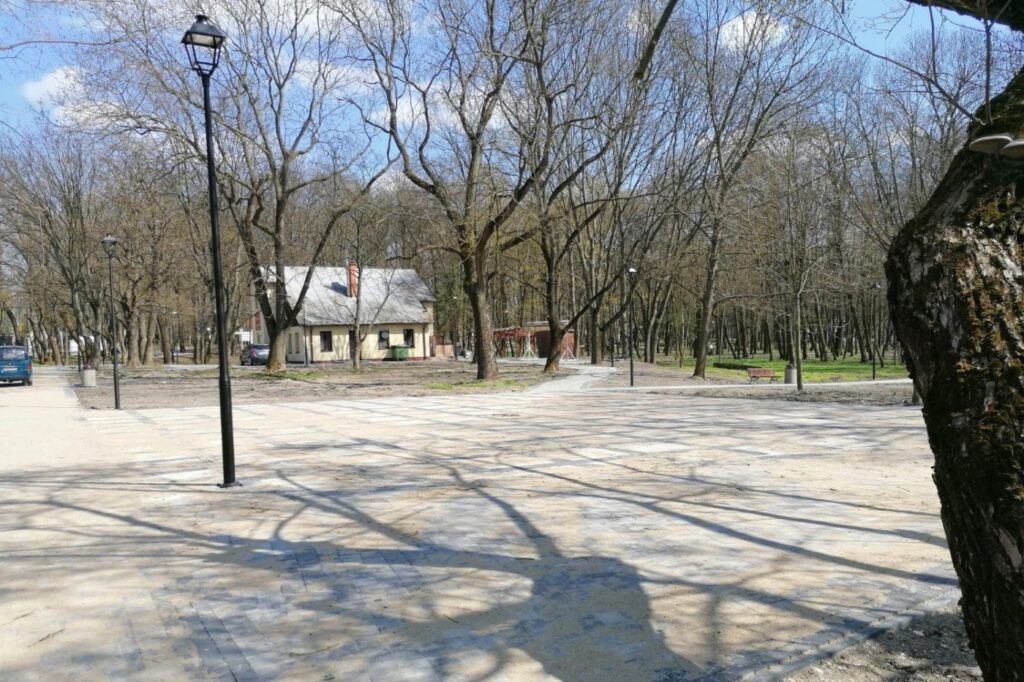 Rozległy plac z jasnych płyt w zadrzewionej części parku, przy zabytkowym parterowym budynku.