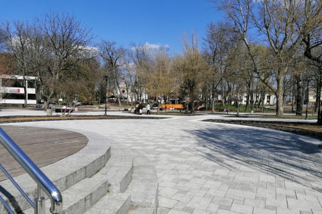Wyniesiony drewniany podest przy placu z szarej kostki, dostępny po trzech stopniach z poręczą.