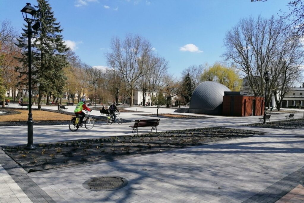 Plac przed muszlą koncertową z równej kostki betonowej, z ławkami i rabatami wzdłuż alejki.