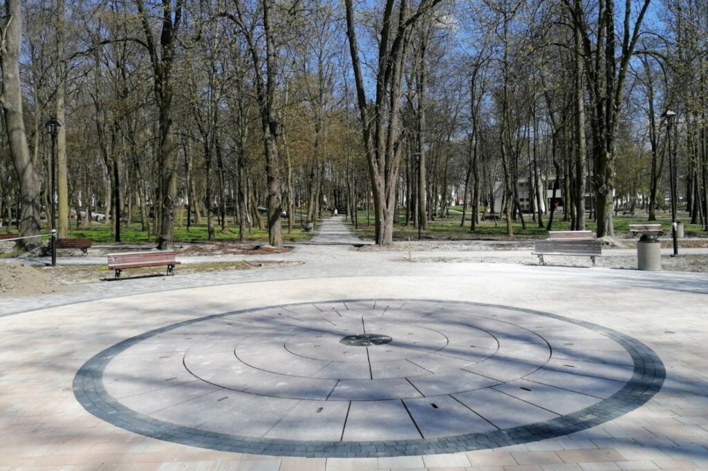 Okrągły plac z koncentrycznym wzorem z kamiennych płyt, w jednym poziomie z alejką, wokół ławki.