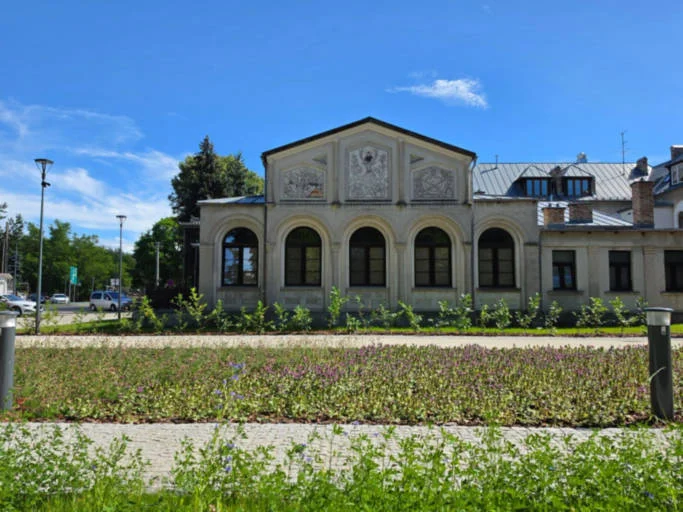 Historyczny budynek dawnego domu kultury z łukowymi oknami, przed nim alejki mineralne i kwitnąca rabata.