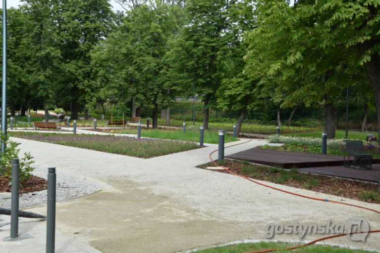 Rozległy plac z jasną nawierzchnią mineralną, niskie lampy i drewniane podesty z krzesłami przy rabatach.