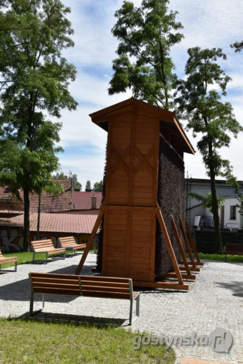 Drewniana tężnia solankowa na placu z drobnej kostki granitowej, dookoła ławki z drewnianymi siedziskami.