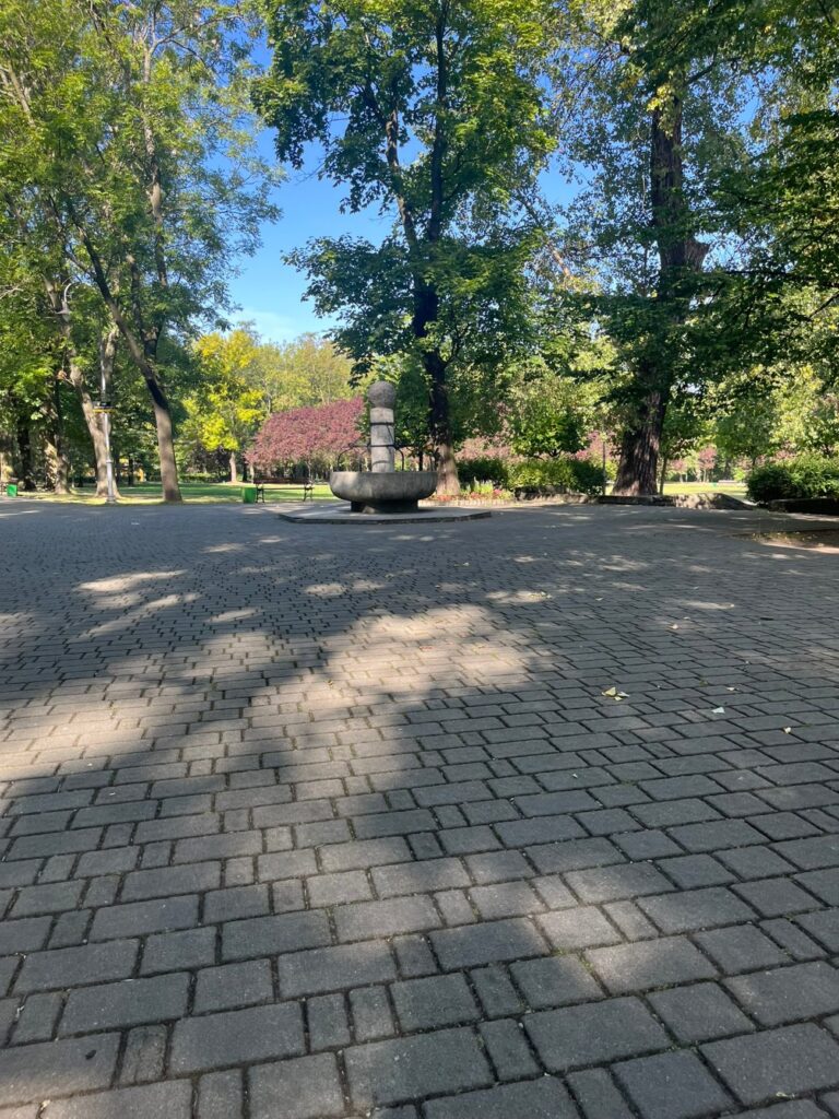 Rozległy plac z kostki brukowej, na skraju kamienna rzeźba, w tle drzewa i kwitnące krzewy.