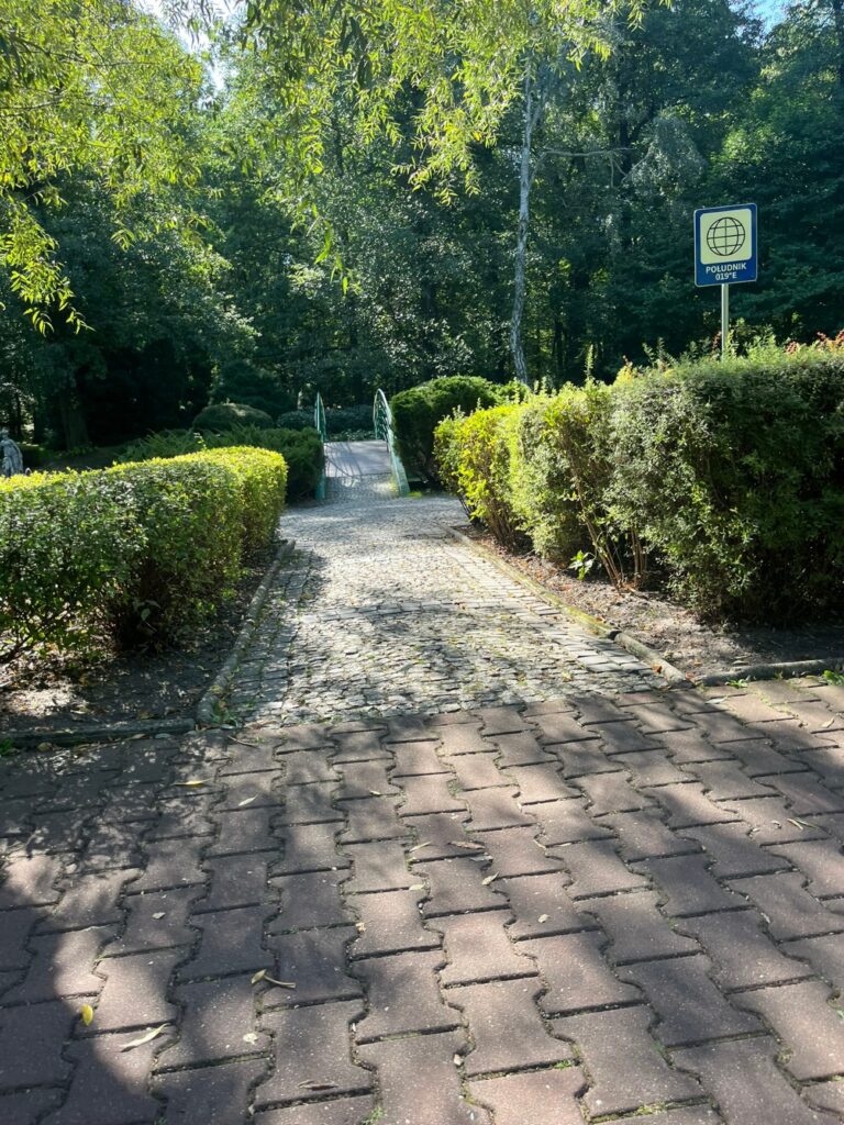 Aleja z kostki brukowej przechodząca w wąską ścieżkę żwirową między strzyżonymi żywopłotami.