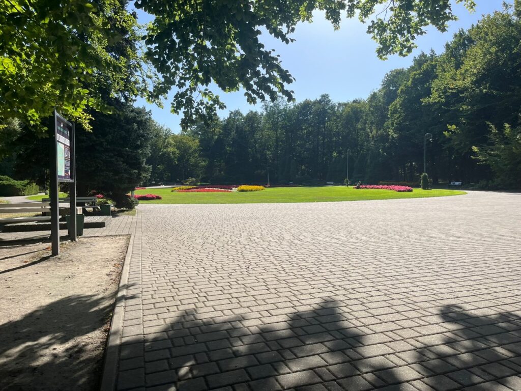 Szeroki plac z kostki brukowej z rzędem ławek i tablicą informacyjną, w tle rabaty kwiatowe i trawnik.