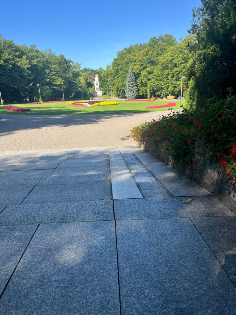 Gładkie, duże płyty granitowe przechodzące w jasną nawierzchnię alei, po prawej murek obsadzony czerwonymi kwiatami.