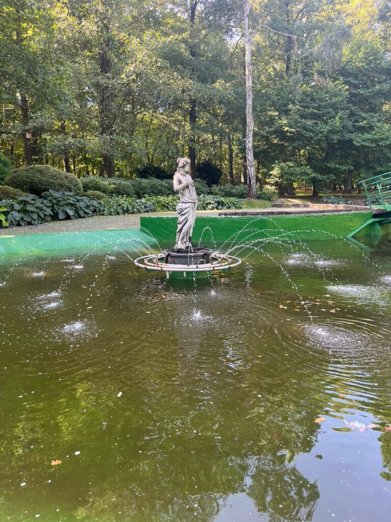 Fontanna z kamienną rzeźbą kobiecej postaci pośrodku wody otoczonej gęstą zielenią.