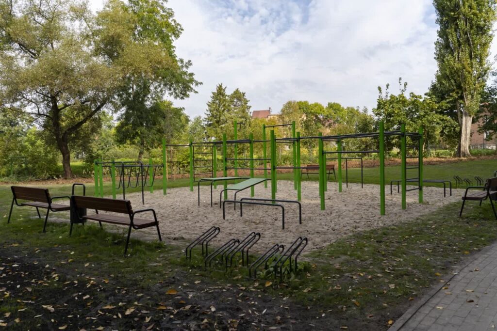 Siłownia plenerowa z zielonymi drążkami na piaszczystym placu, obok ławki, stojak na rowery i alejka z kostki.