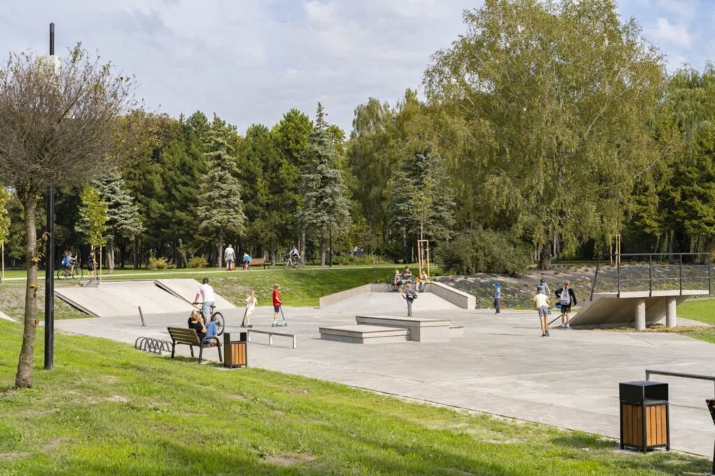 Betonowy skatepark z rampami i murkami u podnóża trawiastej skarpy, przy nim ławka i kosz.