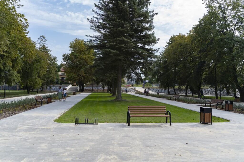Plac z kostki betonowej z ławką i stojakiem na rowery, za trawnikiem dwie alejki prowadzą ku schodom na skraju parku.
