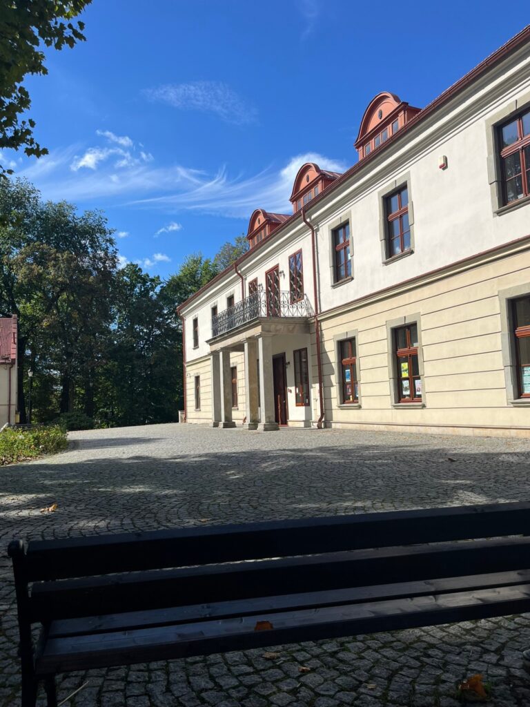 Plac z kostki granitowej przed frontem pałacu z kolumnowym gankiem, na pierwszym planie ławka.