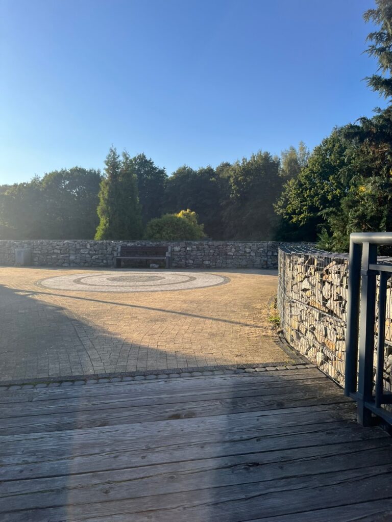 Deski mola przechodzą bez uskoku w plac z beżowej kostki z okrągłym wzorem, przy murku z gabionów ławka.