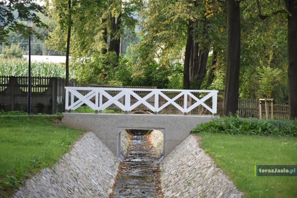 Mostek z białą balustradą nad rowem o brukowanych skarpach, za nim ogrodzenie parku.