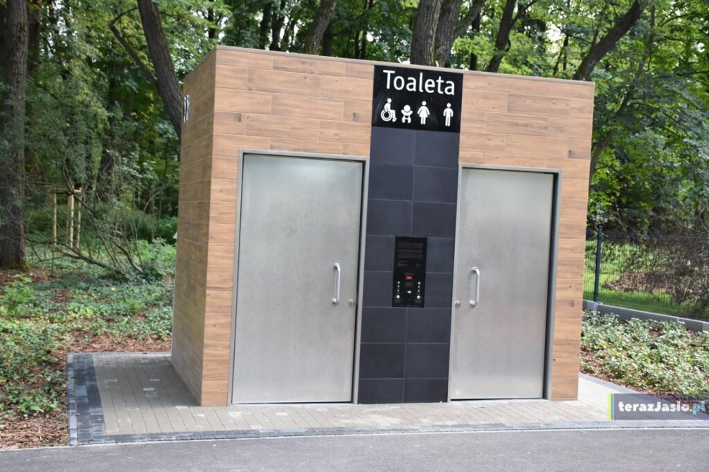 Toaleta publiczna z dwojgiem drzwi, oznaczona dla osób na wózku i z przewijakiem; przed wejściem równa kostka przy alejce.