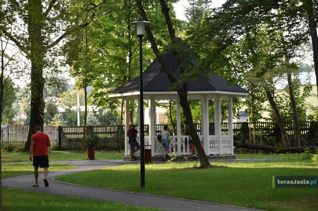 Biała glorieta z ciemnym dachem na niskim podeście przy asfaltowej alejce z latarnią.