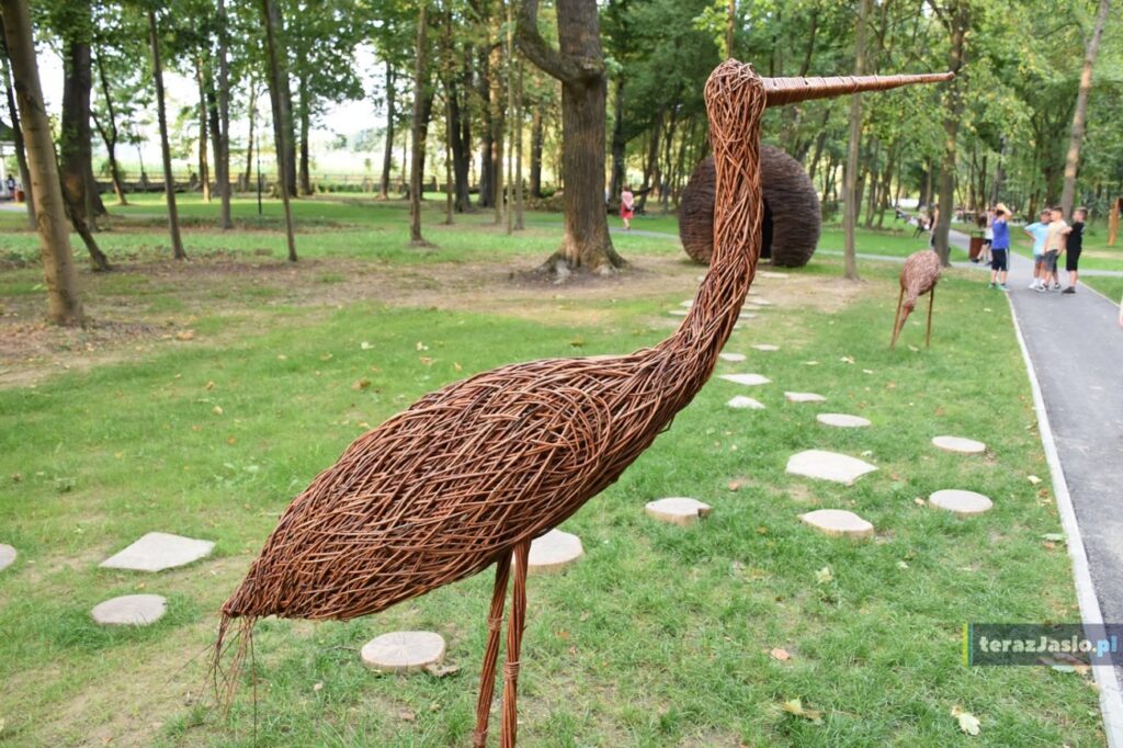 Wiklinowa figura ptaka na trawie, za nią ścieżka z kamiennych płyt i plastrów drewna do wiklinowej chatki.