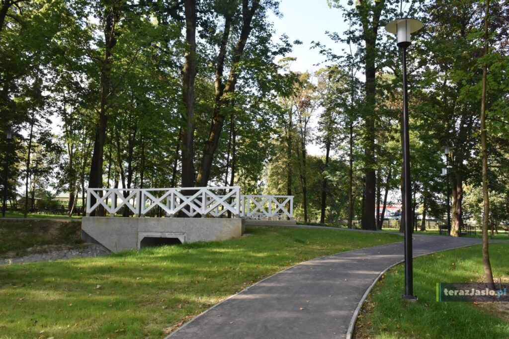 Asfaltowa alejka z latarnią skręca wśród drzew, w tle mostek z białą balustradą nad przepustem.