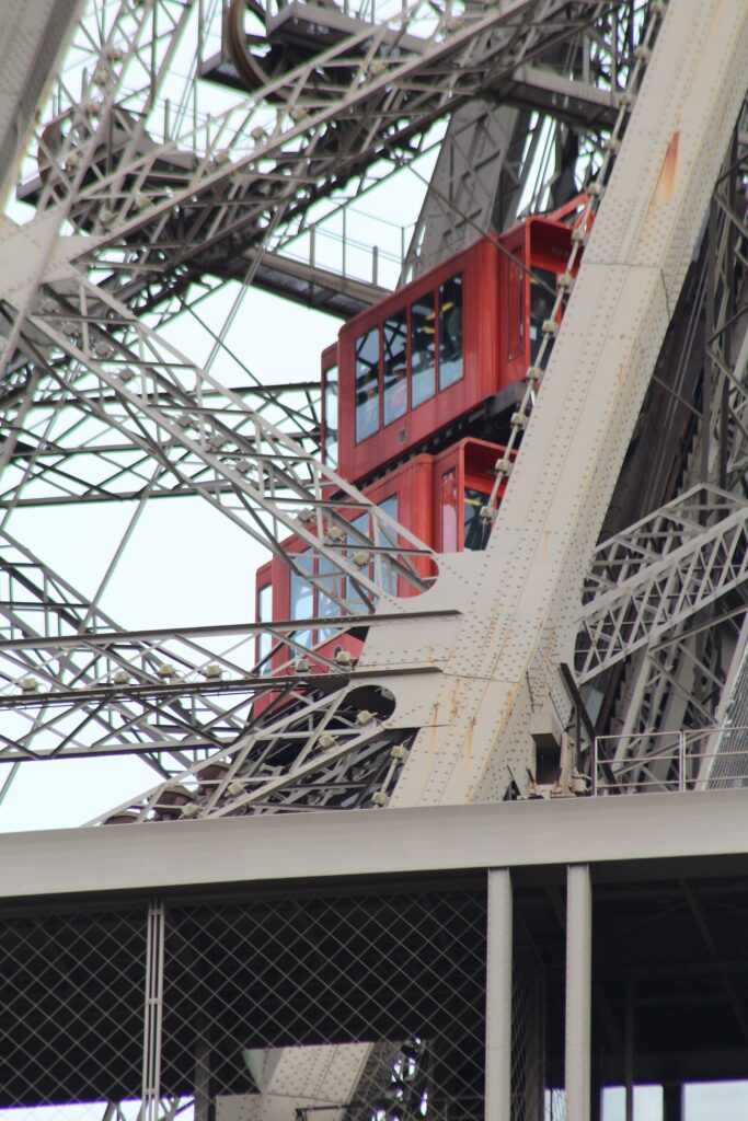 Czerwona kabina windy wjeżdżająca skośnie wzdłuż stalowego filaru Wieży Eiffla